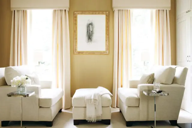 Cream upholstered armchairs flank a cream ottoman in a room with gold textured wallpaper and tall cream curtains framing windows.