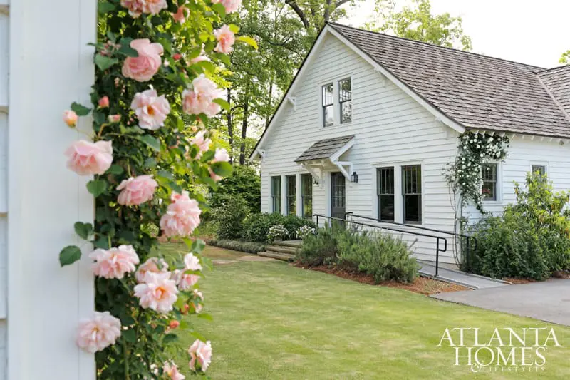 White clapboard house with dark trim windows and a gray shingled roof, surrounded by lush green lawn and flowering rose bushes.