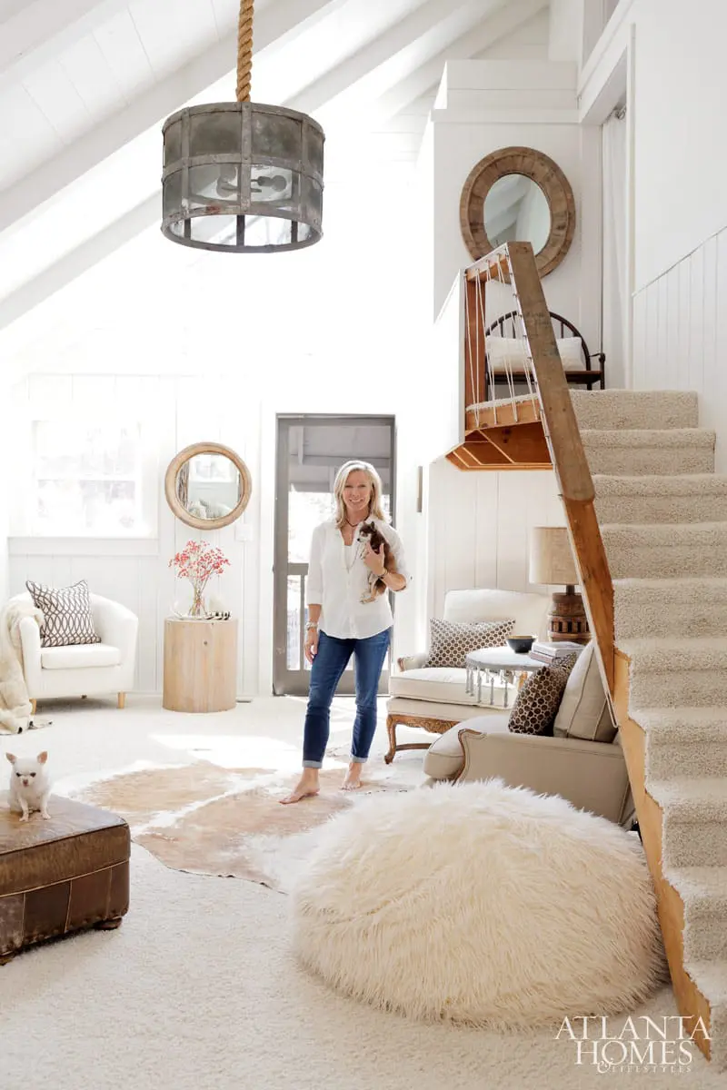 Woman holding a small dog in a bright, airy living room with white walls, vaulted ceiling, and a neutral palette. Features include a cowhide rug, plush ottoman, and a textured pouf.