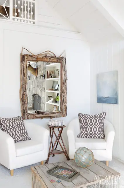 Living room with two white armchairs, a rustic driftwood mirror reflecting a stone fireplace, and a wooden coffee table with a tic-tac-toe game.