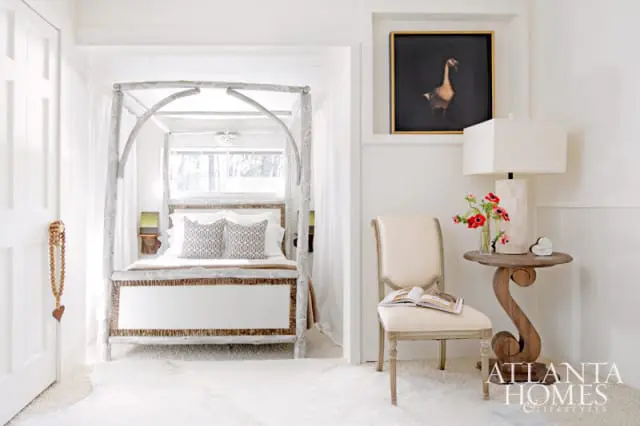 Bedroom with a white canopy bed, upholstered chair, and a side table with a lamp and flowers. Artwork of a goose hangs on the wall.