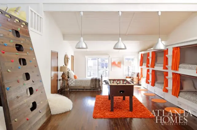Playroom with a wooden climbing wall, foosball table on an orange rug, and built-in bunk beds with orange curtains.