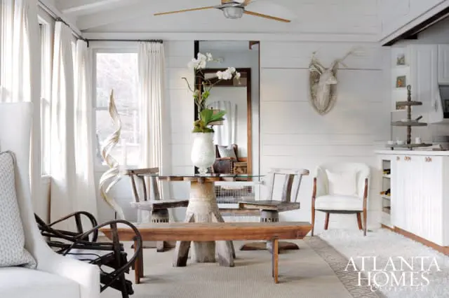 Dining area with glass-top table, rustic wooden chairs and bench, white armchair, and shiplap walls. A mounted deer head adorns the wall.