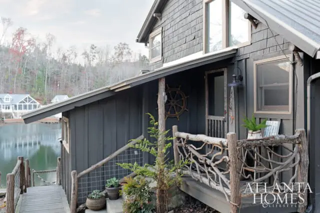 Dark gray wooden house with shingle siding and a rustic porch railing made of branches. A ship's wheel decorates the wall near the entrance. A wooden walkway leads to the water.