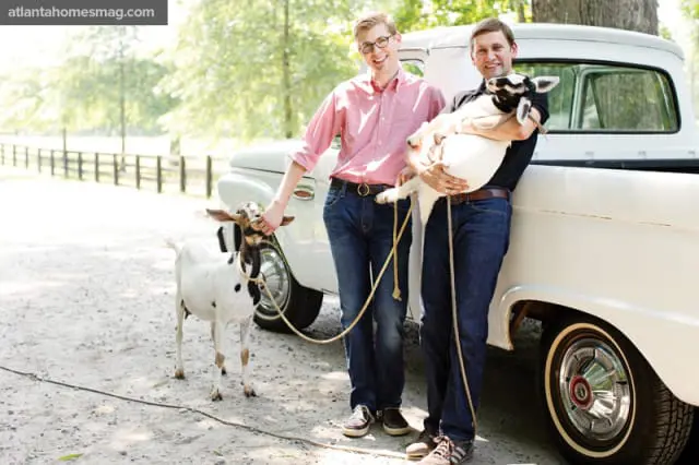 Two men standing next to a white vintage truck with goats. One man holds a goat, the other holds a goat by a rope.