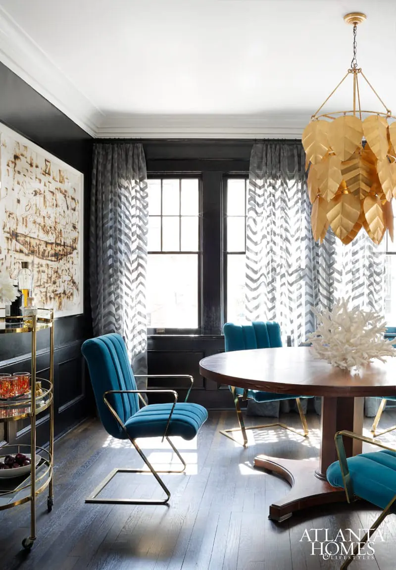 Dining room with dark wood floors, black walls, and teal velvet chairs around a round wooden table. A gold leaf chandelier hangs overhead, and patterned curtains frame the windows.