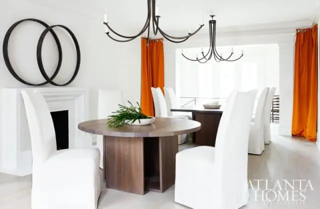 Dining room with white slipcovered chairs, a round wooden table, a dark metal chandelier, and bright orange curtains against a white fireplace
