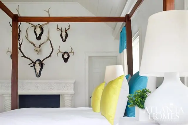Bedroom with a four-poster bed, white fireplace, and mounted deer antlers on a white wall. Yellow and teal pillows accent the bed.
