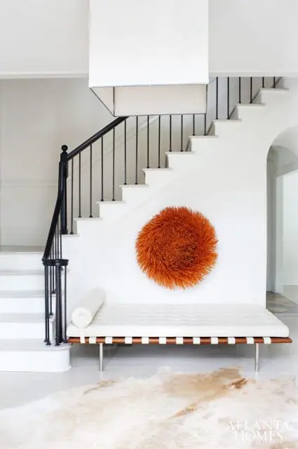 White entryway with a white tufted daybed, a large orange spherical wall art, a black metal staircase, and a light-colored hide rug.