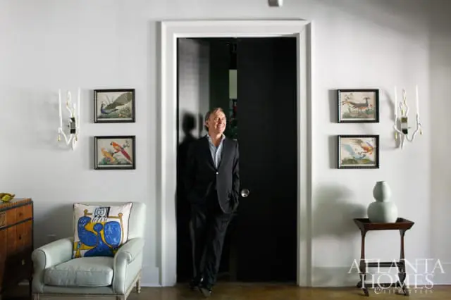 Man in a suit standing in a doorway with black sliding doors, flanked by walls with framed bird art and sconces. A light blue armchair with a colorful abstract pillow sits to the left.