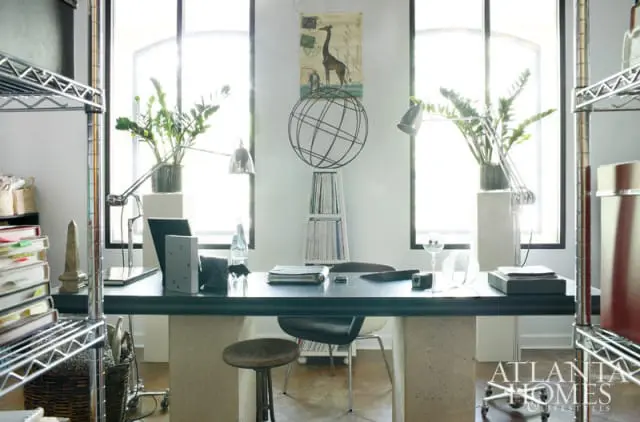 Modern home office with a dark desk supported by concrete pillars, flanked by metal shelving units. Large windows provide natural light, illuminating desk lamps and potted plants.
