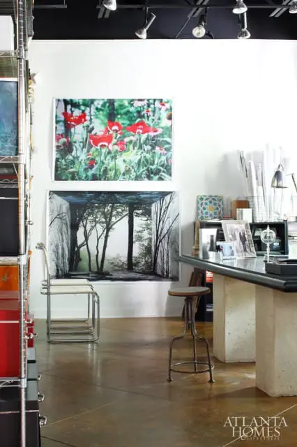 White walls with two large framed artworks: one of red poppies, the other a forest scene. A metal shelving unit with red boxes is on the left. A concrete desk with photos and a lamp is on the right.