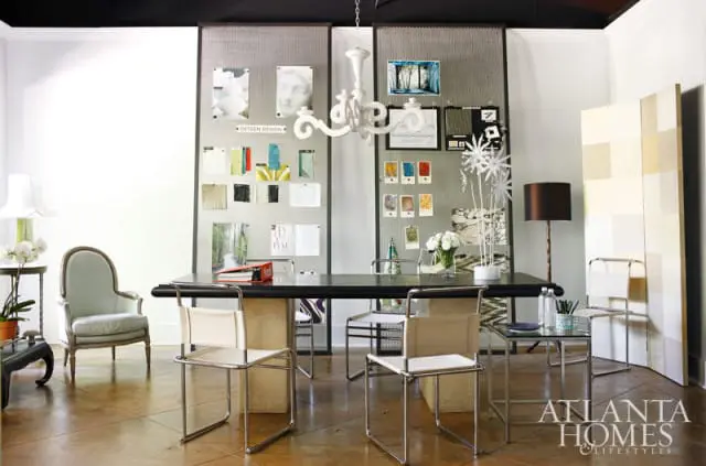 Design studio with a large dark table, chairs, mood boards with fabric swatches and images, and a white chandelier.
