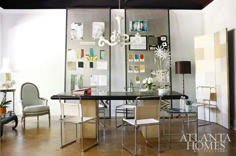 Design studio with a large dark table, chairs, mood boards with fabric swatches and images, and a white chandelier.