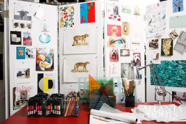 A red desk displays art supplies including markers, colored pencils, stencils, and a desk lamp, with a mood board of images and artwork pinned to white panels behind it.