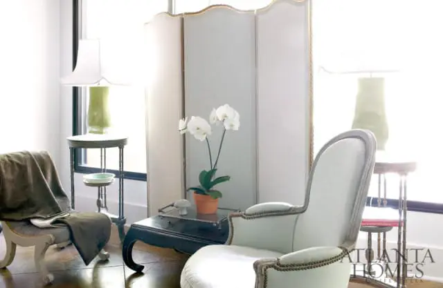 Light blue upholstered armchair with nailhead trim, dark wood coffee table with white orchids, and a cream folding screen in a bright room.