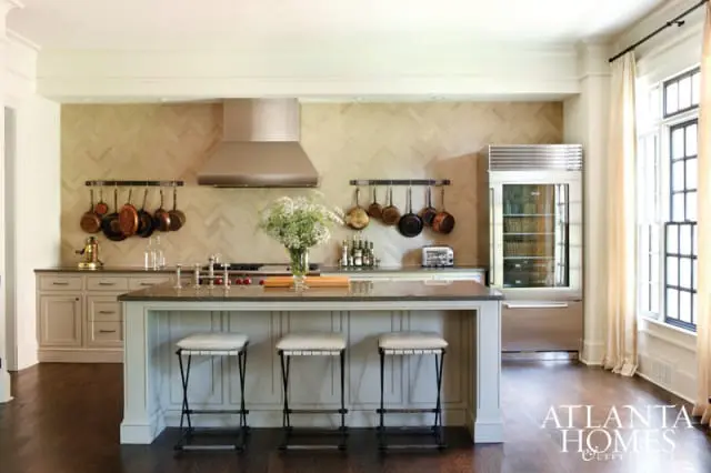 Kitchen with light wood herringbone backsplash, stainless steel range hood, hanging copper pots, and a large island with three stools.