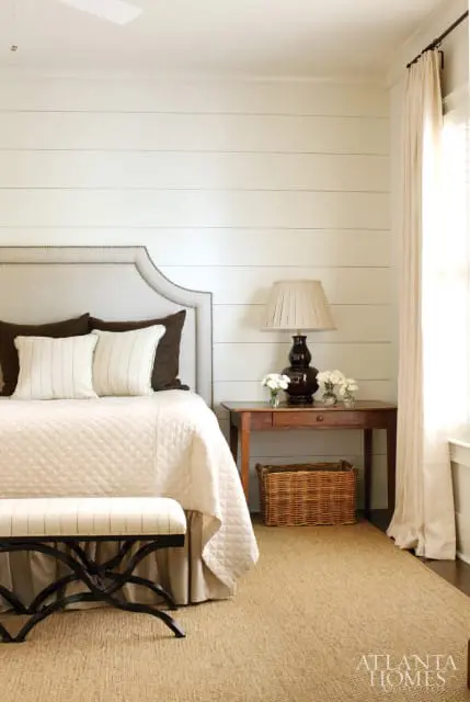 Bedroom with shiplap walls, a tufted headboard, quilted bedspread, and a bedside table with a lamp and flowers.