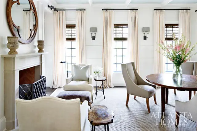 Living room with a stone fireplace, upholstered chairs, a round wooden dining table, and floor-to-ceiling cream curtains.