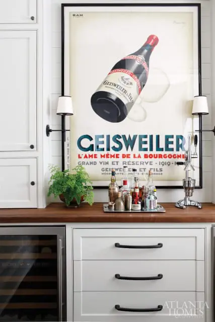 White kitchen bar area with a vintage Geisweiler wine poster, a collection of liquor bottles on a silver tray, a green potted plant, and a chrome citrus juicer.