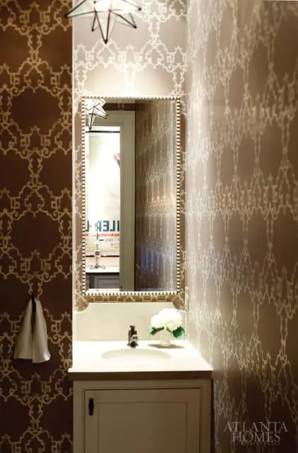 Powder room with brown and beige patterned wallpaper, a vanity with a stone countertop and sink, a decorative mirror, and starburst light fixtures.
