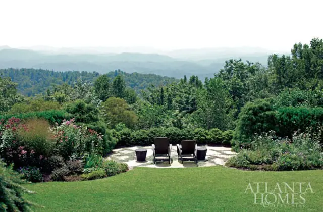 Terraced garden with lush greenery, flowering plants, and two lounge chairs on a stone patio overlooking rolling hills and mountains.