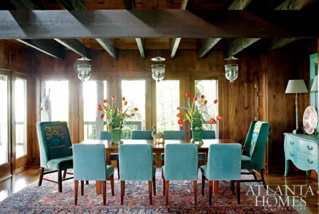 Dining room with wood-paneled walls, exposed beams, and a long wooden table surrounded by teal upholstered chairs. Red tulips in green vases adorn the table.