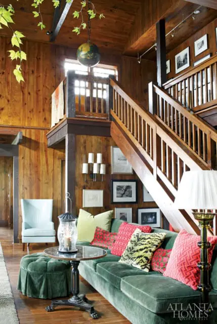 Living room with wood-paneled walls, a green velvet sofa with patterned pillows, and a wooden staircase. A light blue armchair and a green ottoman are also visible.