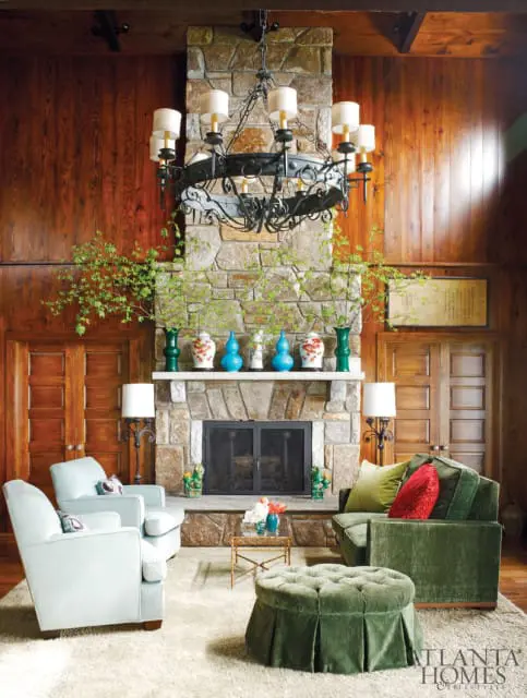 Living room with stone fireplace, wood paneled walls, light blue armchairs, and green velvet sofa and ottoman.