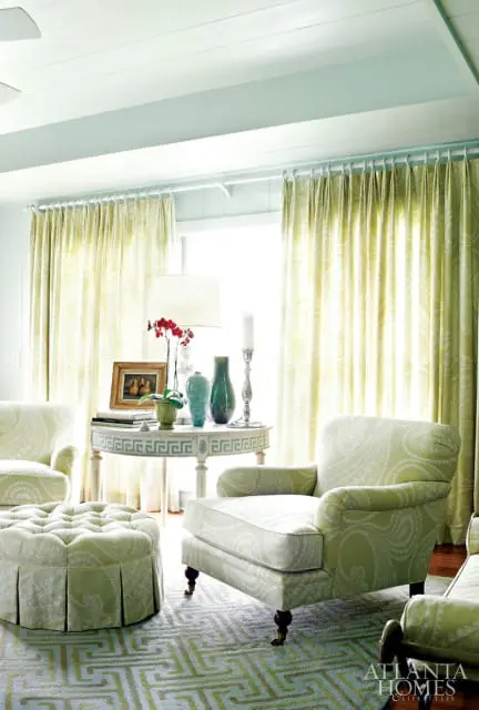 Living room with light blue ceiling and walls, featuring green paisley upholstered armchairs and ottoman, a round table with decorative vases, and floor-to-ceiling curtains.