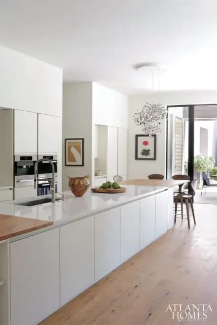 Modern white kitchen with integrated appliances, a large white quartz island with a wooden countertop extension, and a sculptural pendant light.