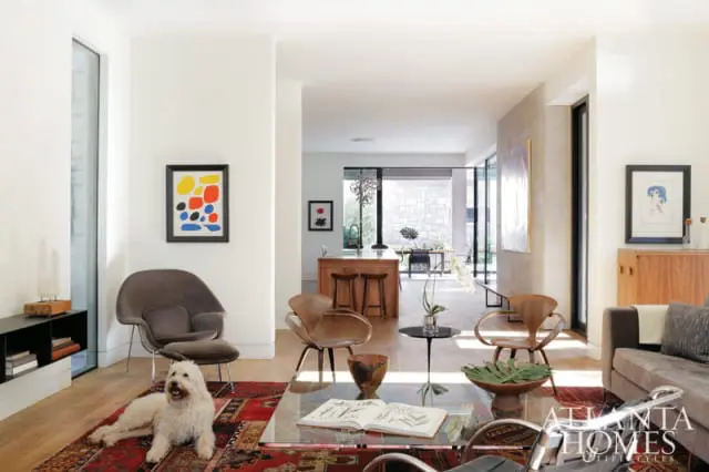 Living room with mid-century modern furniture, abstract art, and a white dog on a Persian rug. A kitchen and dining area are visible in the background.