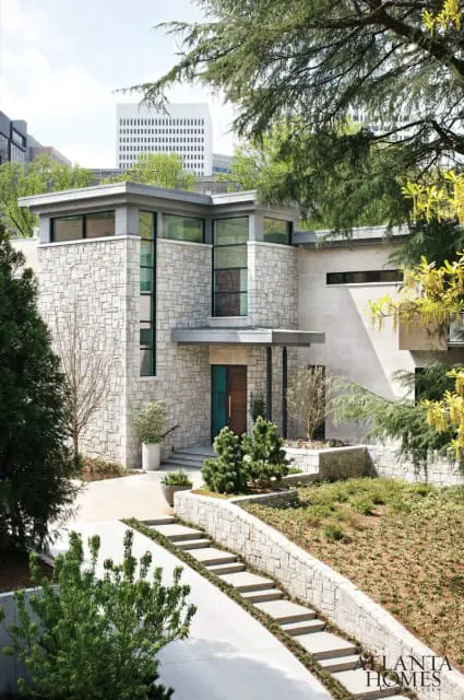 Modern stone house with a flat roof, large windows, and a wooden front door. Stone retaining walls and concrete steps lead to the entrance.