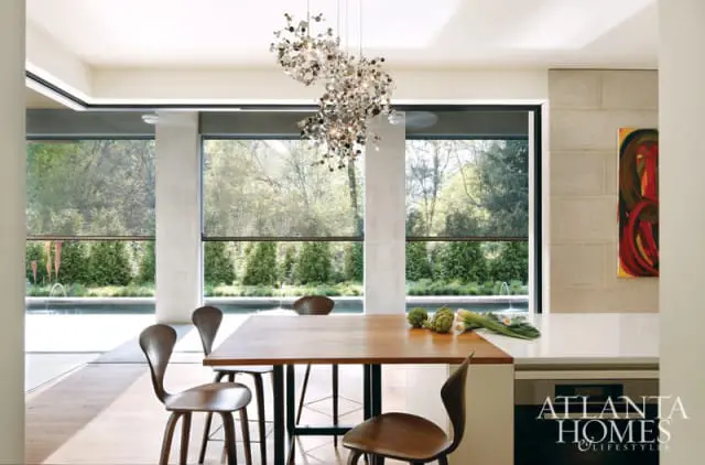 Dining area with a wooden table and chairs, featuring a modern chandelier and large windows overlooking a pool and garden.