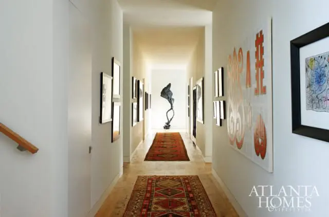 Hallway with light gray walls, wood floors, and two Persian runners. Artwork adorns the walls, and a sculpture stands at the far end.