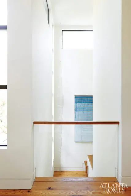 Modern hallway with white walls, glass and wood railings, and a blue abstract painting. Natural light streams in from large windows.