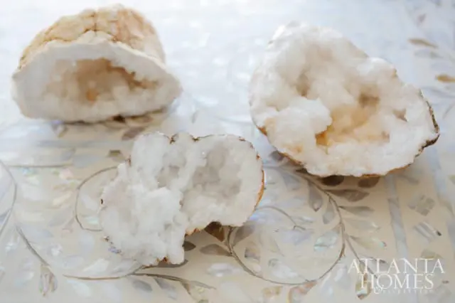 Three halved geodes with white crystal interiors resting on a decorative surface with a floral inlay pattern. The Atlanta Homes & Lifestyles logo is visible in the lower right corner.