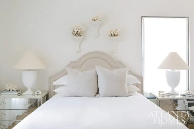 Serene bedroom with a tufted upholstered headboard, crisp white bedding, and neutral linen pillows. White coral accents adorn wall shelves above the bed. Mirrored nightstands flank the bed, each holding a white lamp.