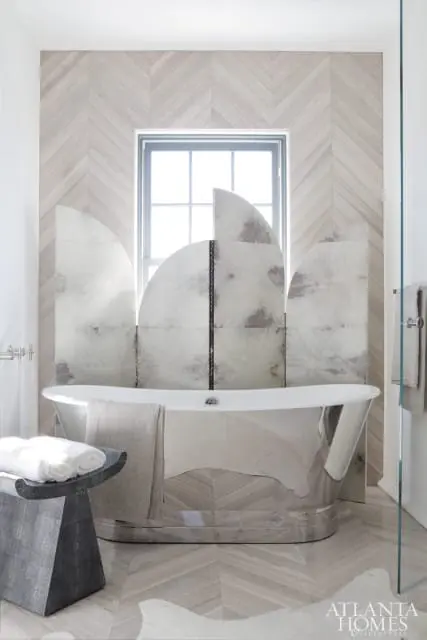 Modern bathroom with a freestanding silver bathtub, a dark gray sculptural stool with stacked white towels, and a decorative screen with arched panels behind the tub.