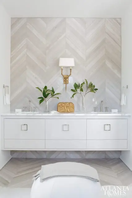 Modern bathroom with a white floating vanity, marble countertop, and a wall clad in light gray chevron tile. A gold sconce illuminates the space above two sinks.