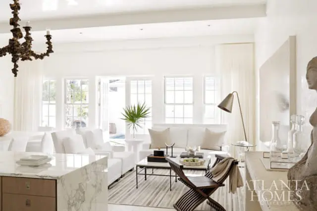 Bright living room with white sofas, marble island, striped rug, and ornate chandelier. Large windows and doors open to an outdoor space.
