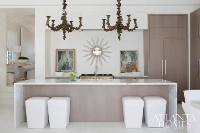 Modern kitchen with a marble waterfall island, taupe wood cabinetry, dual ornate chandeliers, and a sunburst mirror above the range.