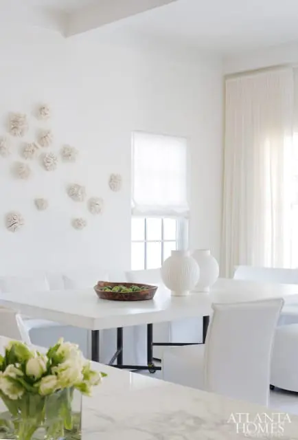 White dining room with a white rectangular table, white chairs, and white textured wall art. A wooden bowl of green fruit sits on the table between two white spherical vases.