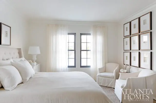 Cream bedroom with tufted headboard, white bedding, sheer curtains, two upholstered armchairs, and a gallery wall of framed art.