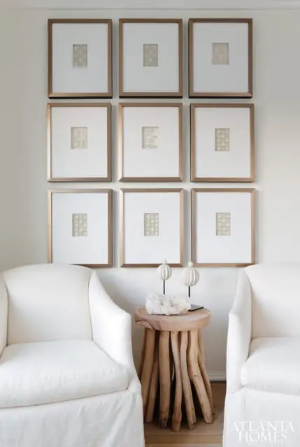 Two white slipcovered armchairs flank a rustic wooden side table topped with a crystal geode and two decorative spheres. Nine framed artworks with a grid pattern hang on the wall above.