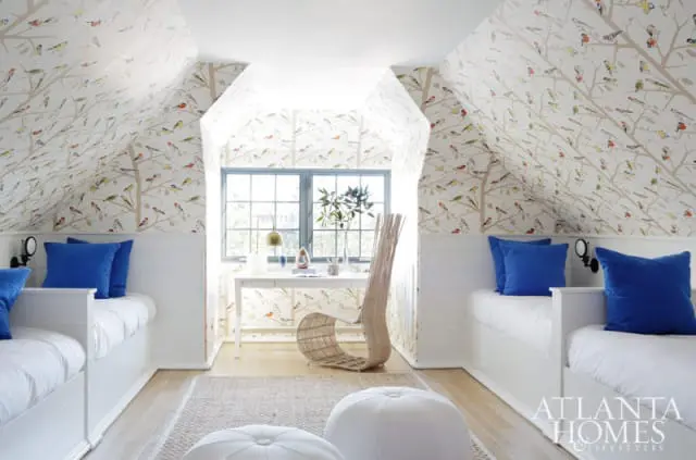Attic bedroom with bird-themed wallpaper, white shiplap walls, twin beds with blue pillows, a desk with a wicker chair, and a jute rug.