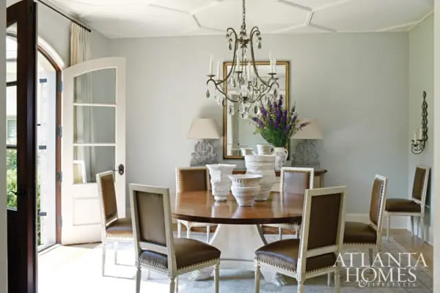 Dining room with a round wooden table, brown leather chairs with white trim, and a crystal chandelier. A large mirror hangs above a buffet with decorative vases and lamps.