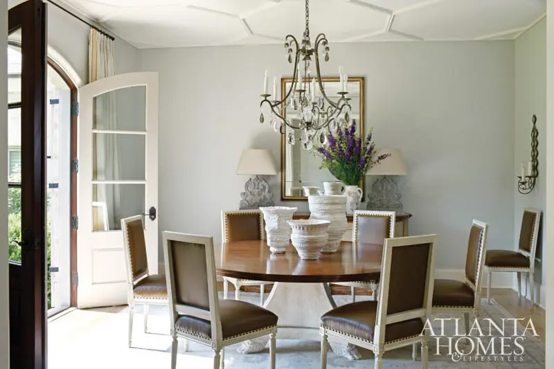 Dining room with a round wooden table, brown leather chairs with white trim, and a crystal chandelier. A large mirror hangs above a buffet with decorative vases and lamps.