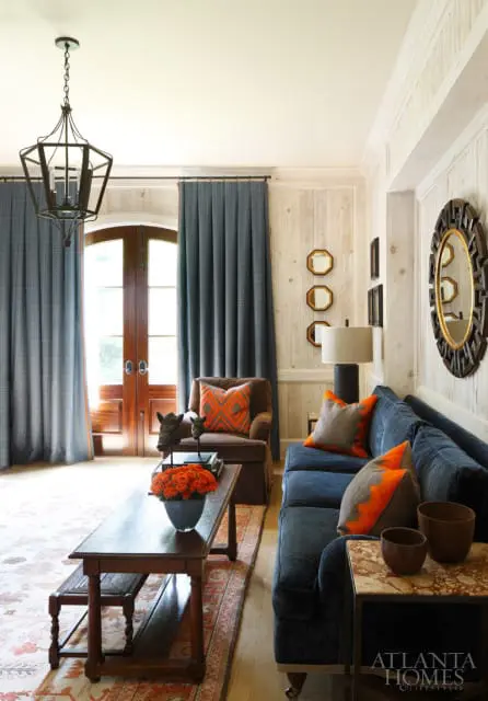 Living room with navy blue sofa, brown armchair, wood paneled walls, dark wood coffee table with orange roses, and a large geometric mirror above a side table.