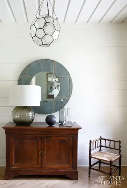 Wooden cabinet with glass lamp, decorative spheres, and bottles below a round distressed metal mirror and geometric pendant light.